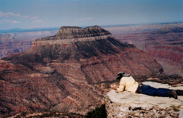 Person lying on cliff edge sketching a distant flat-topped mesa