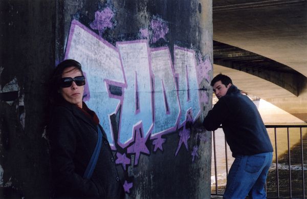 Two people stand by a graffiti-covered bridge pillar near water