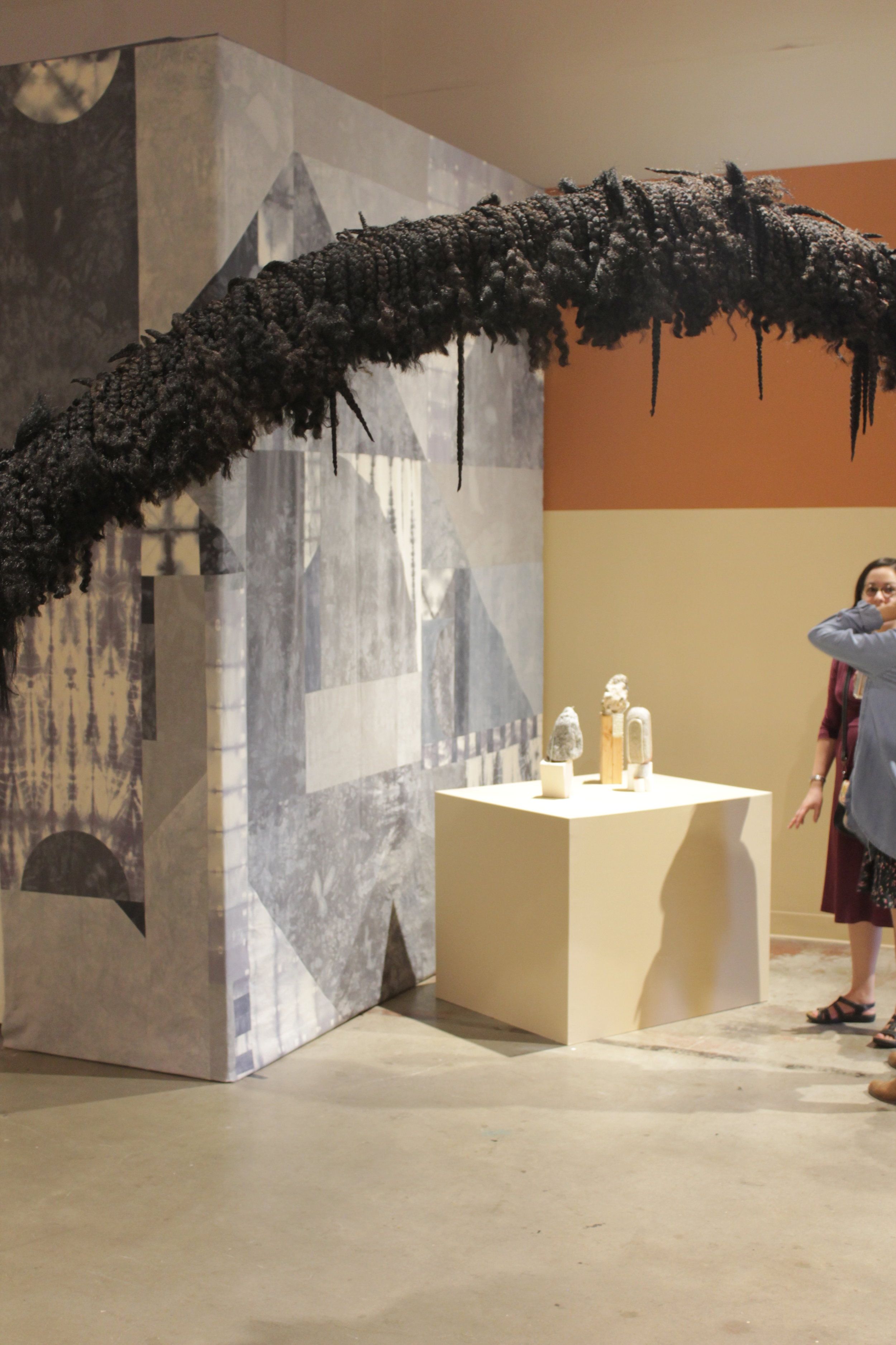 Art installation with arching braided structure above pedestal of small sculptures