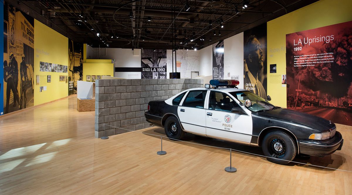 Exhibition view including police car from 1992, brick wall, large scale photos, and wall text