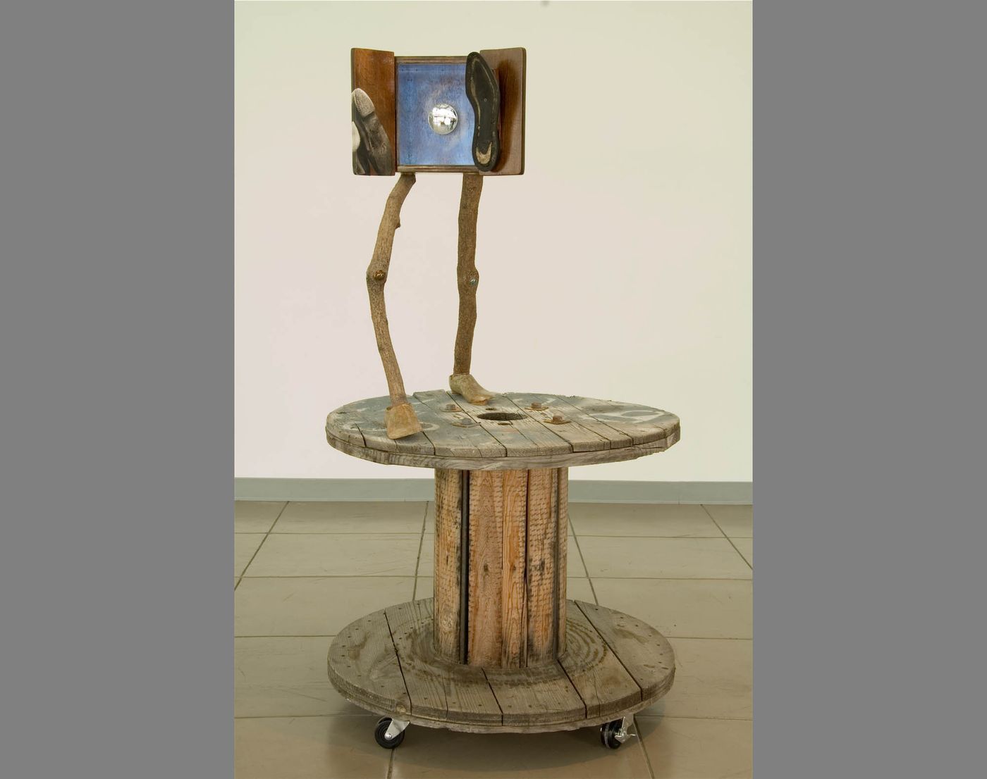 Mixed media assemblage with humanoid limbs atop industrial wood cable spool