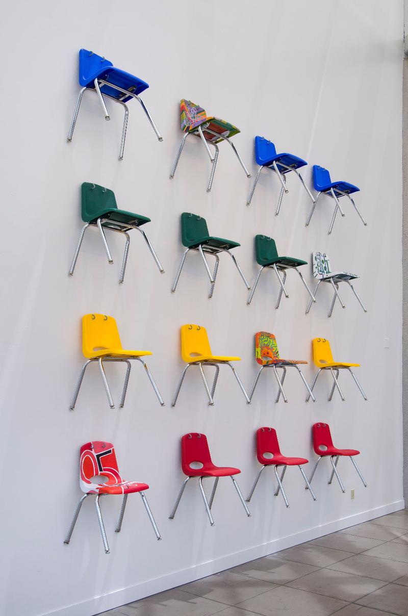 Installation of 16 school chairs in blue, green, yellow, and orange all mounted on the wall in a grid