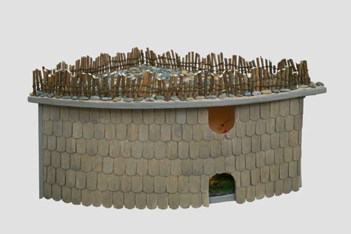 Maquette with shingled exterior; arched window and door; wood fence lining roof