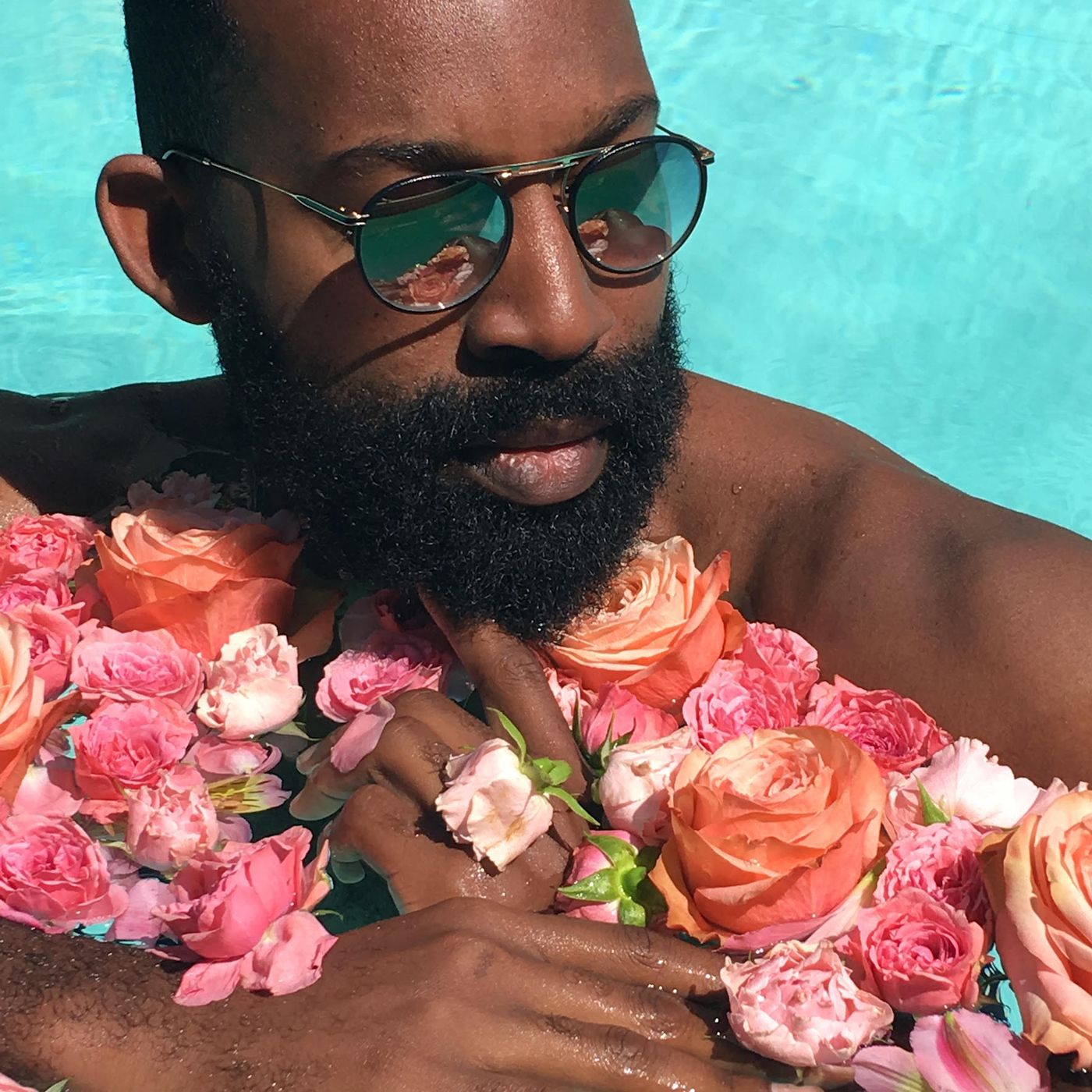 Bearded Black man in mirrored sunglasses standing in aqua colored pool holding dozens of orange and pink flowers