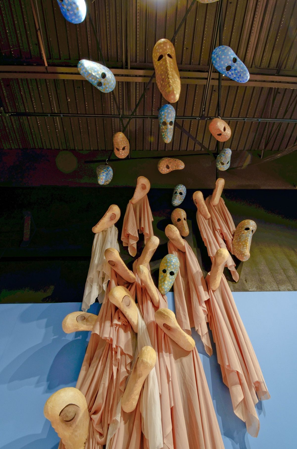 Ethereal assemblage of putty and blue colored disembodied heads, masks, and faces installed from ceiling
