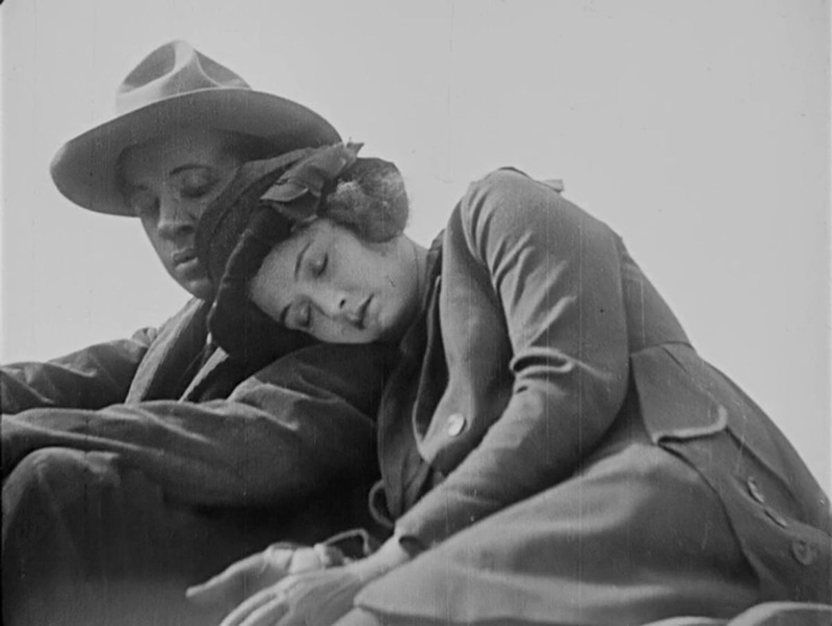 Black and white still showing woman asleep at a man's shoulder, they are dressed for travel