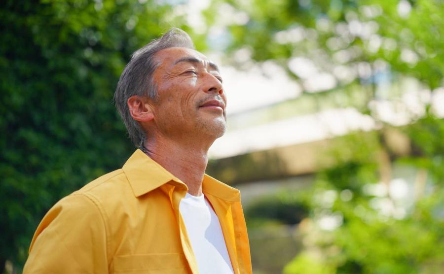 An older East Asian man in a yellow shirt tilts his head back with eyes closed outside, taking a deep breath and looking fulfilled as he considers taking on an encore career.