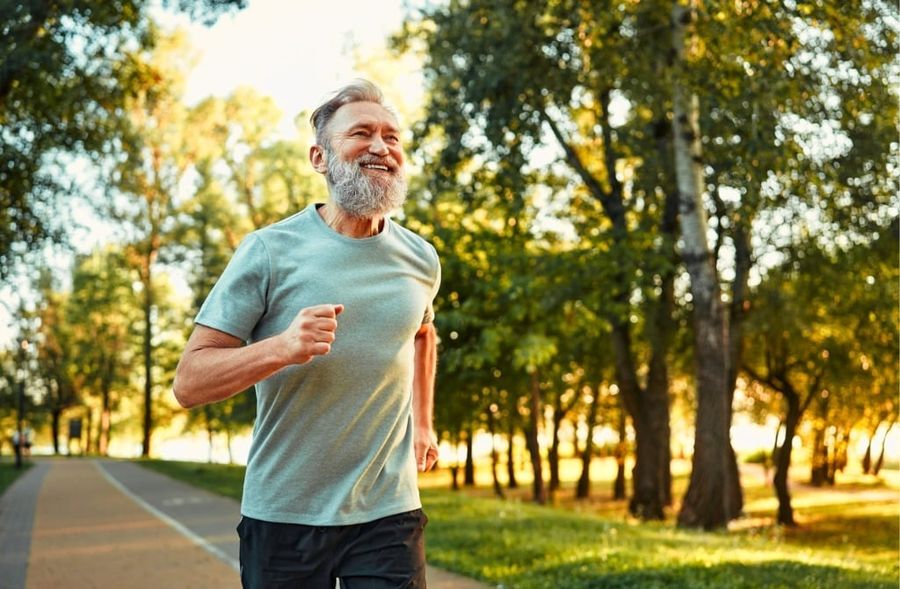 A smiling, bearded senior man jogs along a paved path in a sunny park lined with trees.