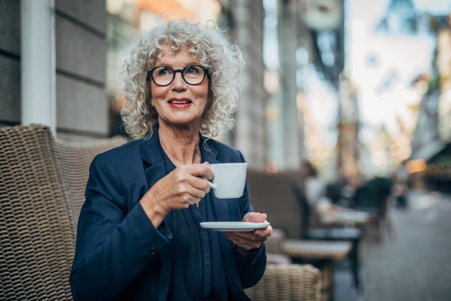 A woman with curly gray hair and glasses sits outdoors, drinking coffee from a small white cup and saucer.