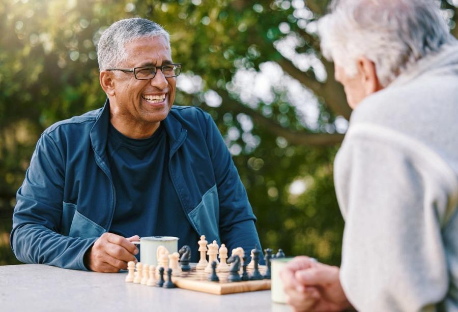 Two older men share a laugh while playing chess outdoors, showing how shared hobbies offer a natural answer for how to make friends as an adult.