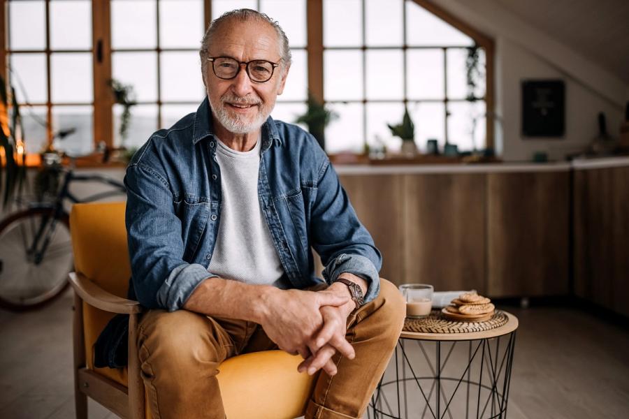 A stylish, confident senior man in a denim shirt sits comfortably in a chair at home, reflecting how modern retirees stay informed on emerging personal finance trends.