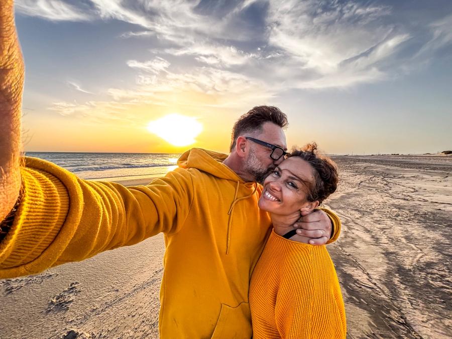 A couple taking a selfie on a sandy beach at sunset as the man kisses the woman's cheek, celebrating their new freedom during the transition to retirement.