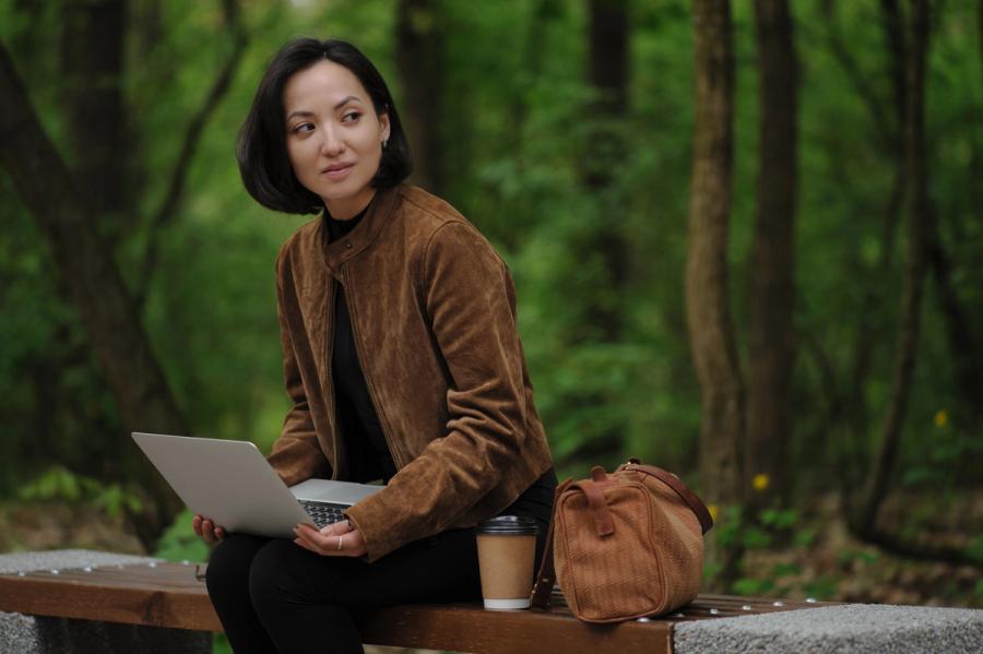 A woman sitting on a wooden park bench with an open laptop and a coffee cup, surrounded by trees.