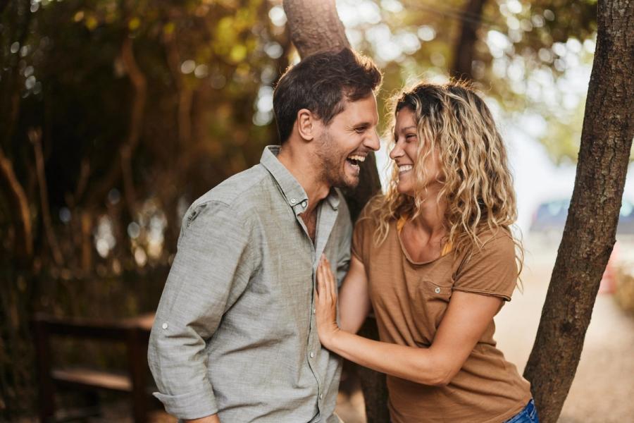 A laughing couple stands outdoors near trees, laughing and looking at each other.