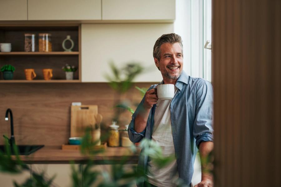A smiling mature man stands in a modern kitchen enjoying a cup of coffee while looking out the window, reflecting the contentment of adhering to solid personal finance rules.