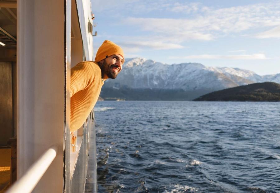 A smiling man in a yellow beanie and sweater leans out from a boat deck window, looking across open water toward snow-capped mountains.