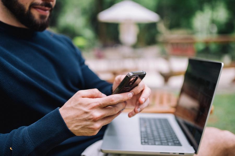 Young man working remotely in the garden, sitting on an outdoors chair and using cell phone and laptop