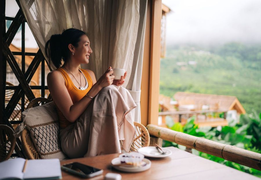 A smiling woman rests on a wicker chair with a mug on a balcony overlooking lush, foggy mountains, capturing a quiet moment of personal freedom and how how wealth is defined beyond just money.