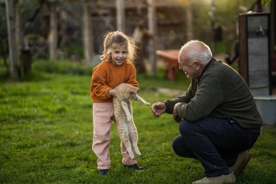 A smiling young girl in an orange sweater holds an orange tabby cat while her grandfather kneels beside her in a yard.
