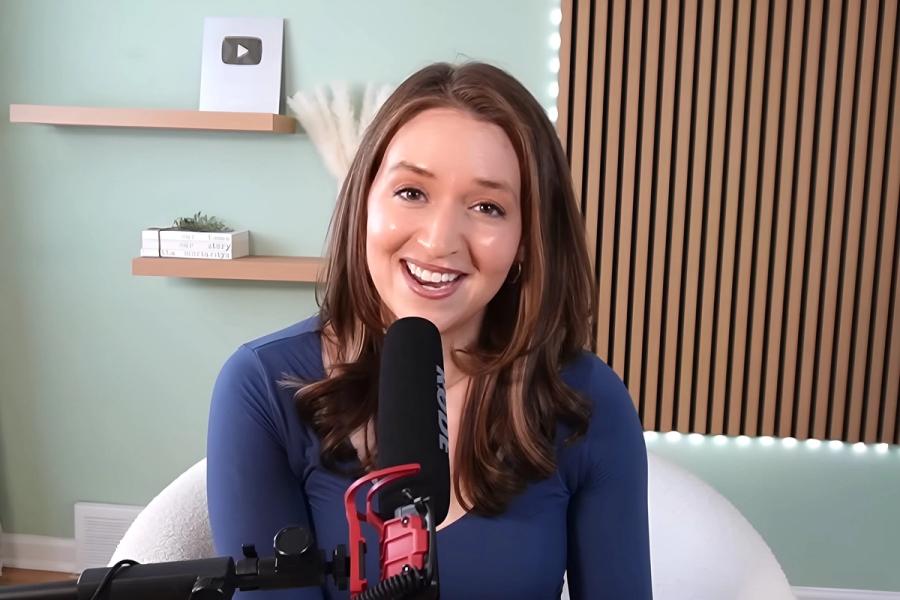 Erin Moriarty, the creator of Erin Talks Money, smiles warmly while speaking into a microphone during a video recording in her studio, featuring a modern backdrop with wooden slats and floating shelves.