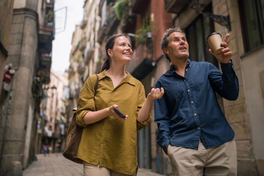 A man pointing into the distance with a coffee cup while walking down a narrow European street alongside a woman holding a phone.