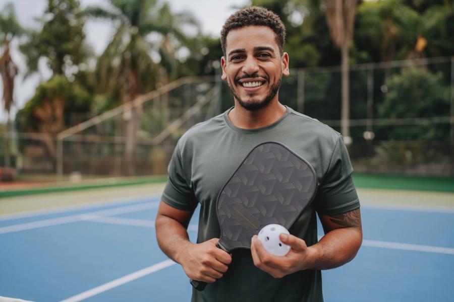 A smiling man holds a pickleball paddle and ball while standing on an outdoor court.
