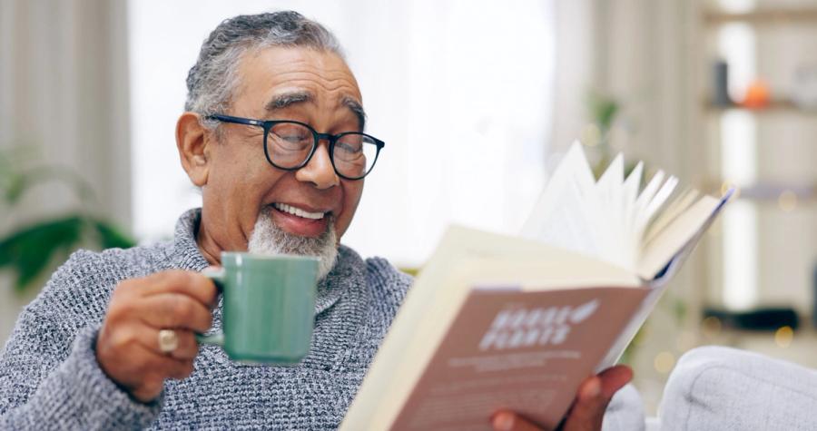 A smiling older man in glasses and a gray sweater holds a green mug while reading a book at home.