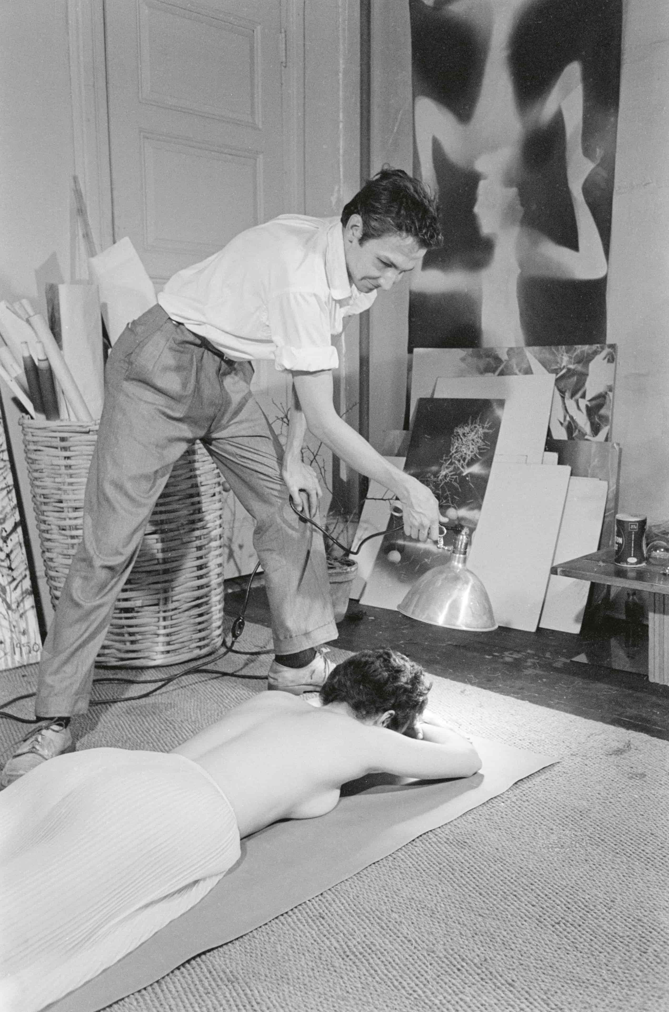 A black-and-white photograph depicting a person using a lamp to provide light or warmth to a woman lying face down on a mat, surrounded by art supplies and artworks in a studio setting.