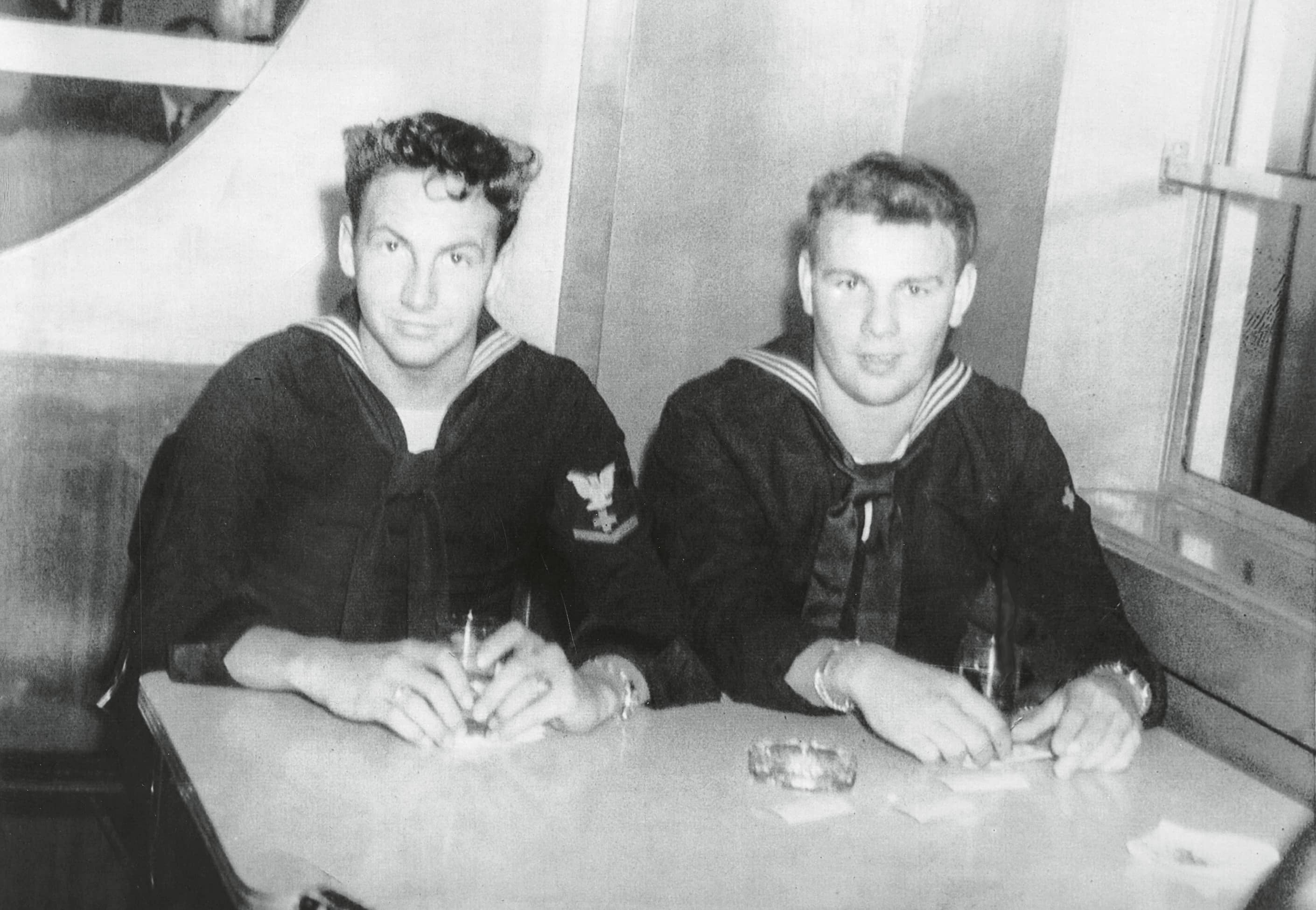 Two young men in naval uniforms sit at a table, each holding a drink, with a cigarette pack and ashtray visible in front of them.