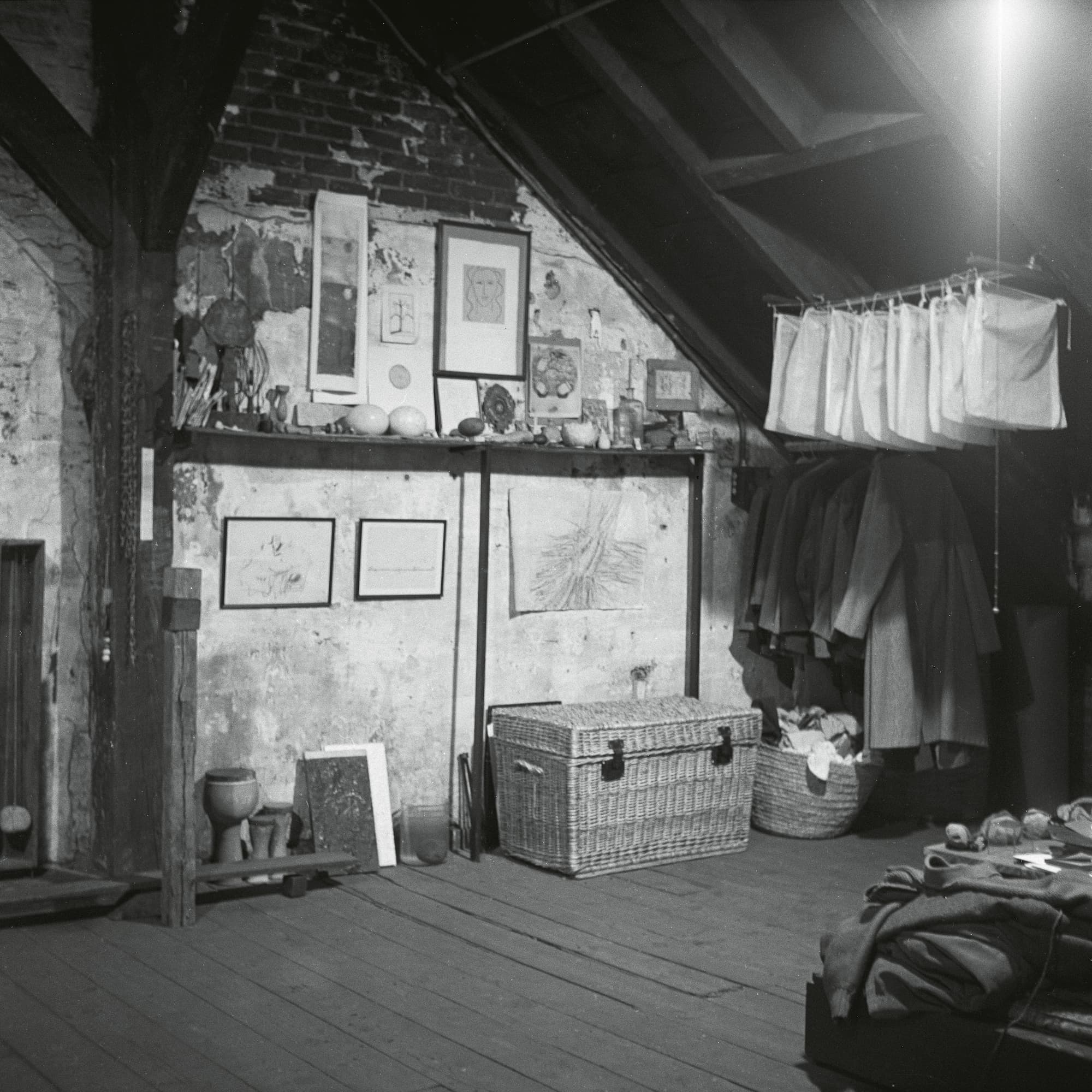A dimly lit attic space featuring wooden beams, shelves adorned with various artworks and objects, a wicker basket, and hanging garments.