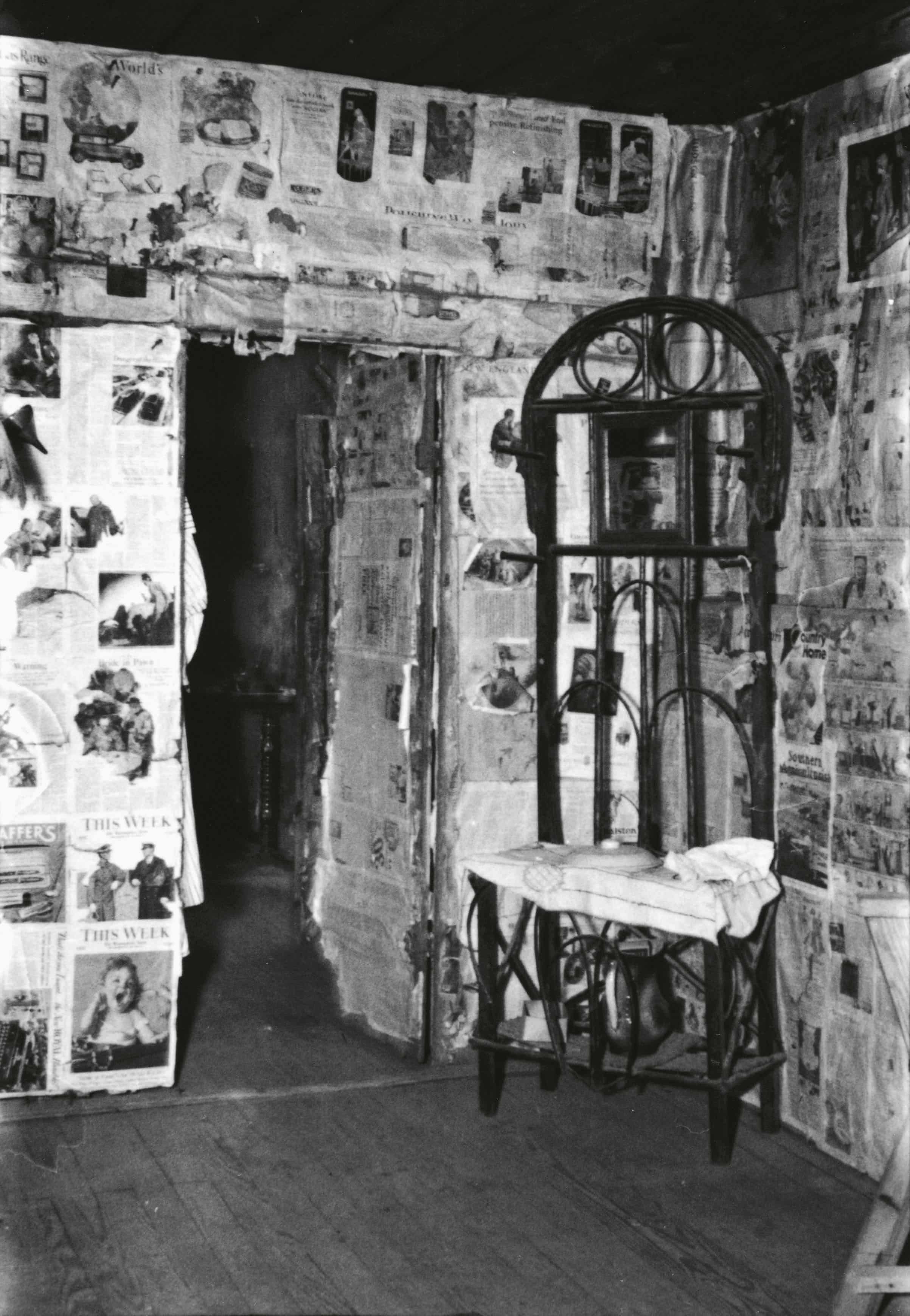 A black and white photograph depicts a small, sparsely furnished room with walls covered in pasted newspaper clippings, featuring a tall, ornate mirror and a small table.