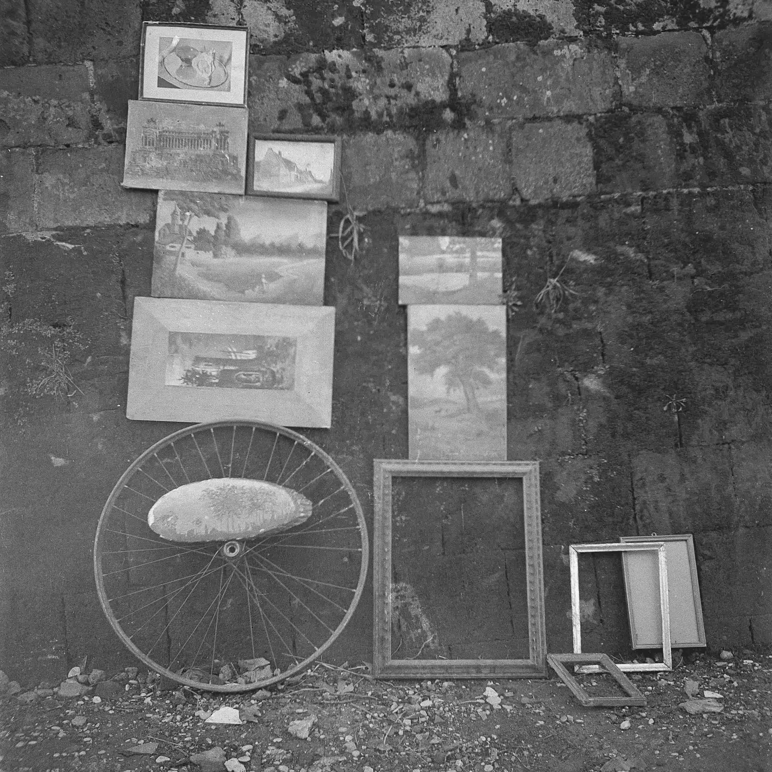 A collection of framed paintings and empty picture frames leaning against a textured stone wall, with a bicycle wheel resting on the ground beside them.