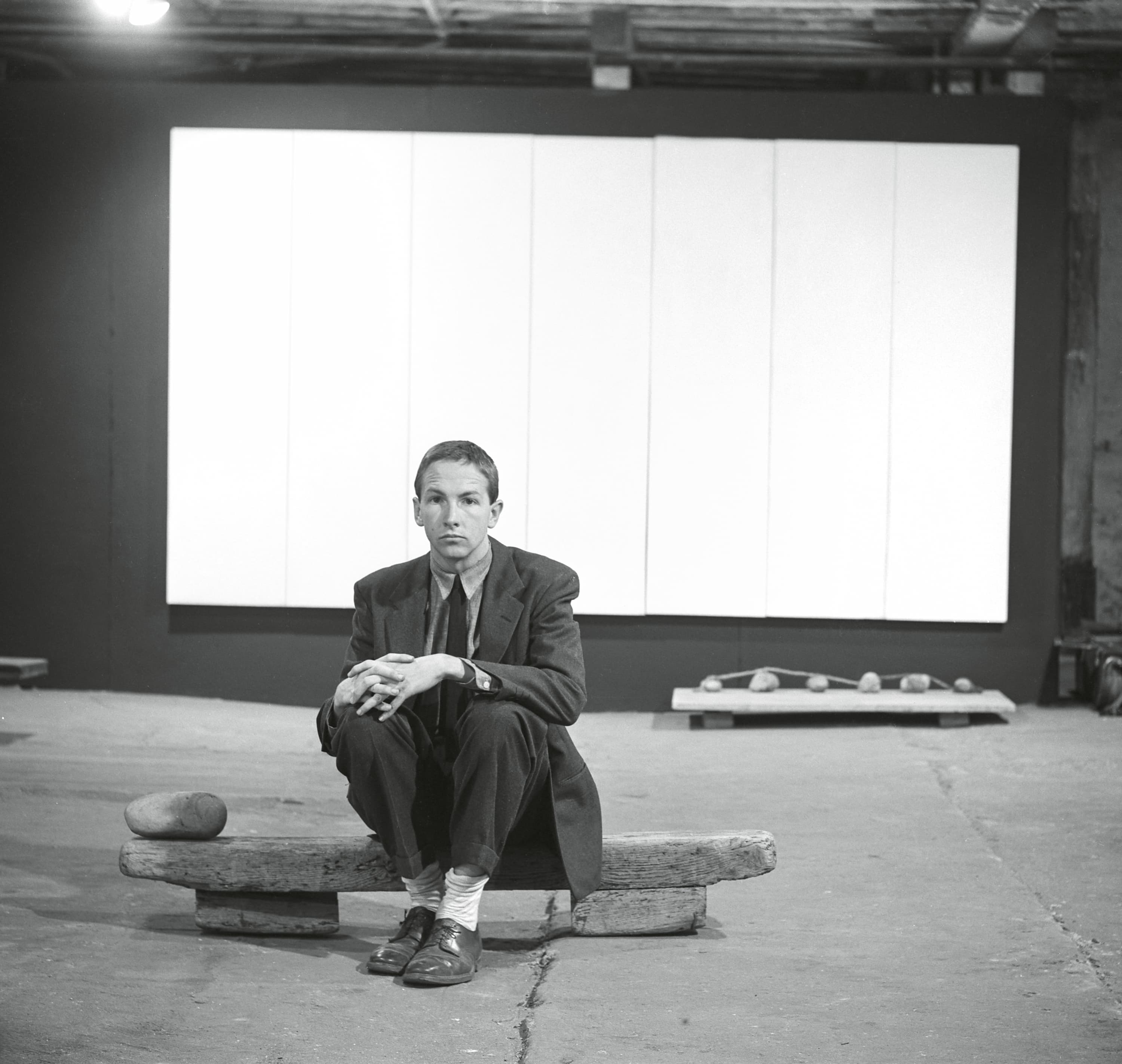 A person in a suit sits on a wooden beam in an art space, with a large blank canvas and a few stones visible in the background.