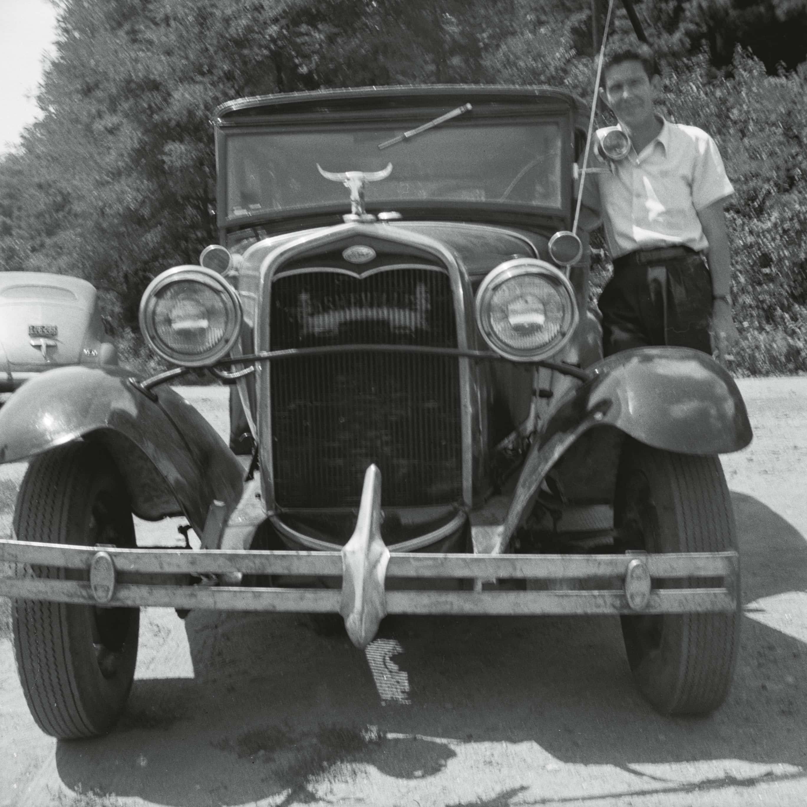 A man stands beside a vintage car from the 1920s, showcasing its distinctive front grille and hood ornament, with trees in the background.
