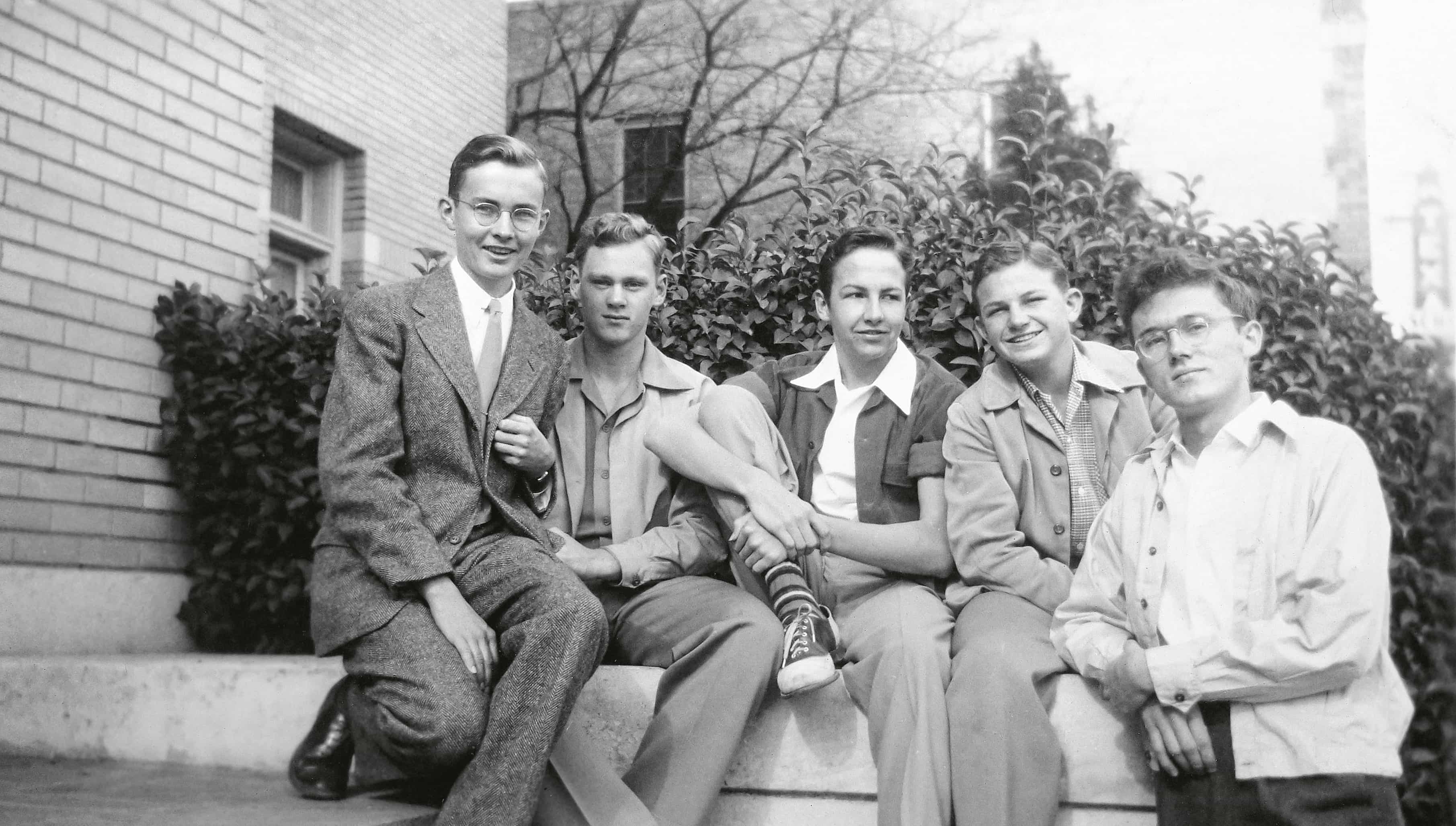 A group of five young men, dressed in 1940s attire, sit together on a stone ledge in front of a brick building, smiling and posing for the camera.