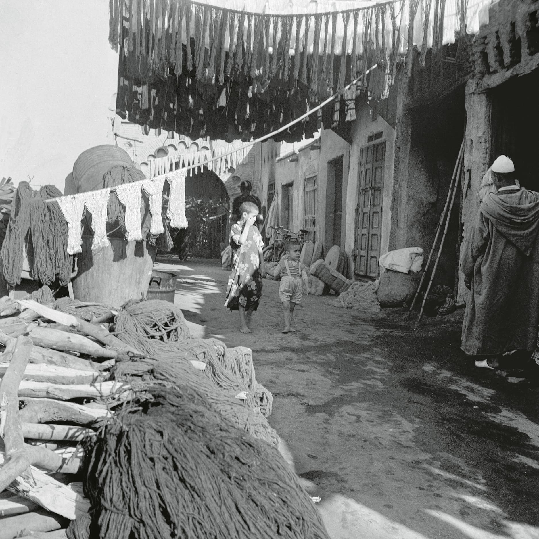 A black-and-white photograph of a narrow alleyway lined with hanging textiles, where a woman and a child walk past stacks of dyed materials, while a figure in traditional attire observes from the side.