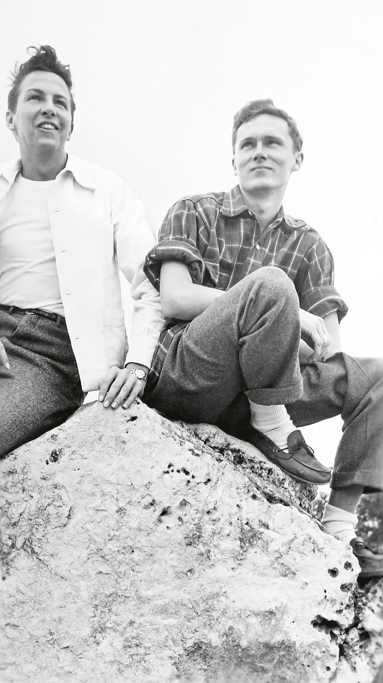Two young men sit on a large rock, one wearing a white shirt and the other in a plaid shirt, both looking thoughtfully into the distance.