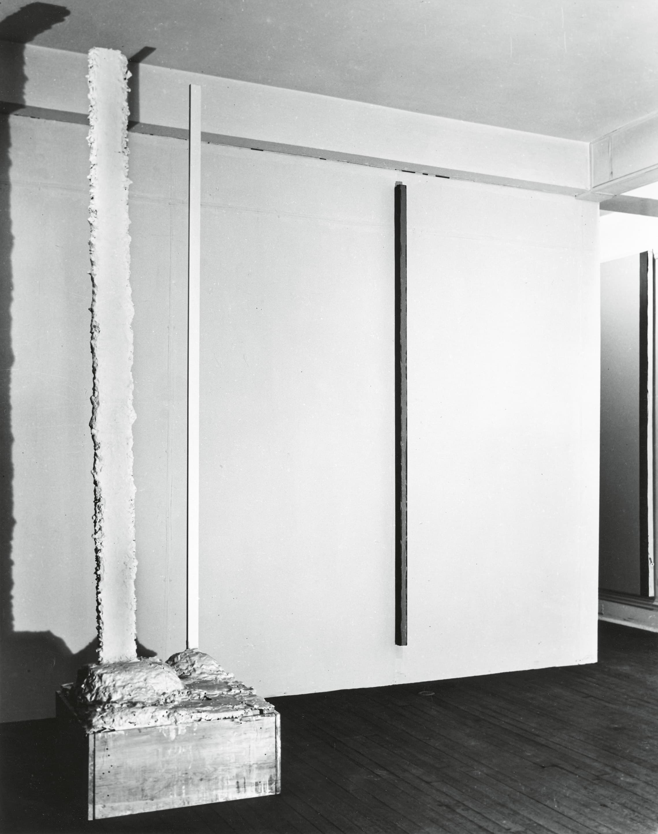 The image shows a minimalist art installation featuring a tall, textured vertical sculpture on a pedestal next to two slender, smooth vertical bars mounted on a plain wall.