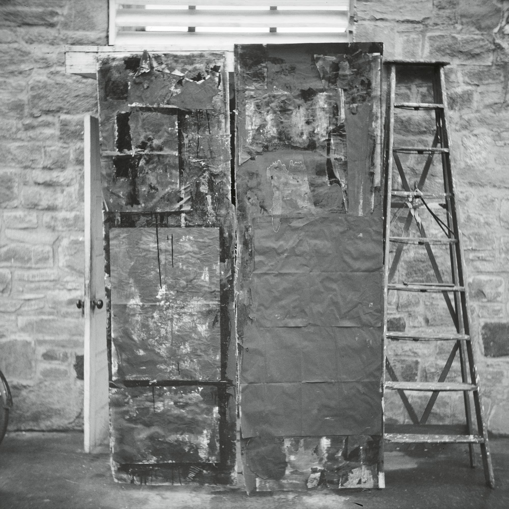 A black and white photograph of a textured, abstract wall covered in paint and materials, with a wooden ladder leaning against it in a rustic setting.