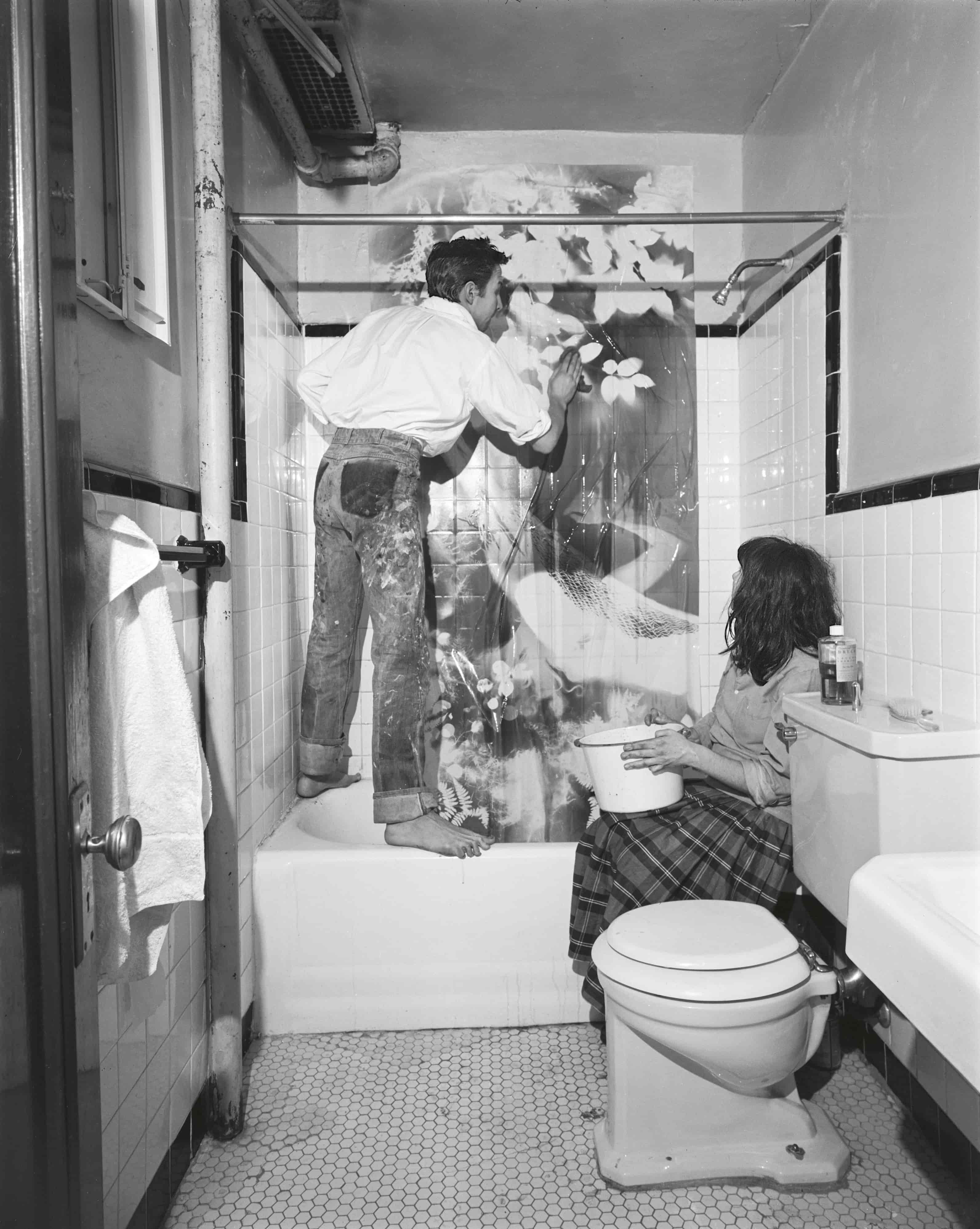 A man stands on the edge of a bathtub, painting a mural on the wall, while a woman sits on the floor nearby, holding a bucket and looking up at him in a vintage bathroom setting.