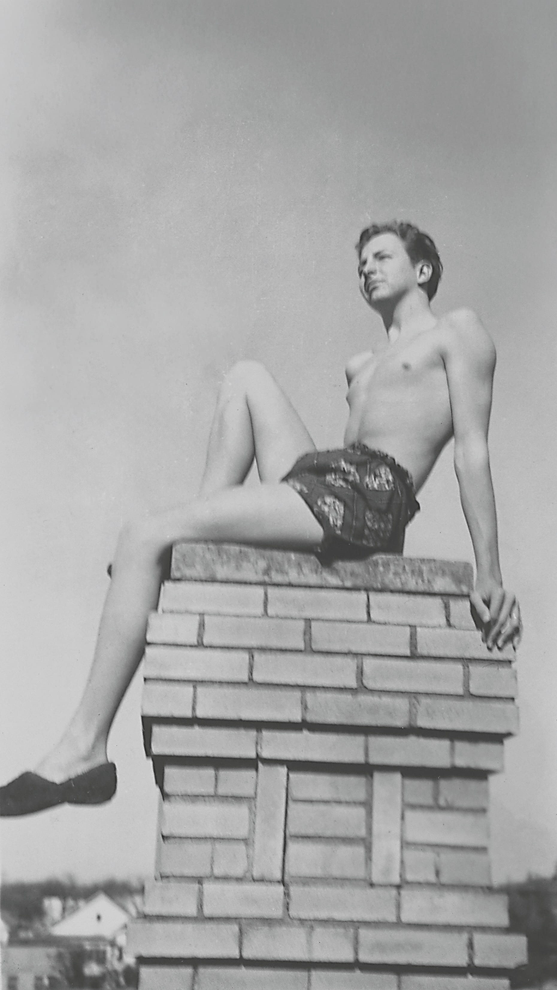 A young man sits casually on a brick chimney, wearing shorts and black shoes, with a clear sky in the background.