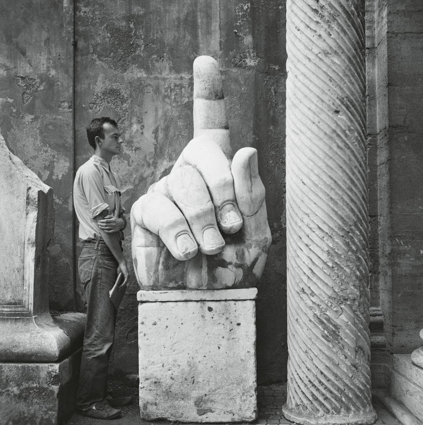 A man stands beside a large, ancient stone sculpture of a hand with an extended finger, set against a backdrop of weathered stone columns and walls.