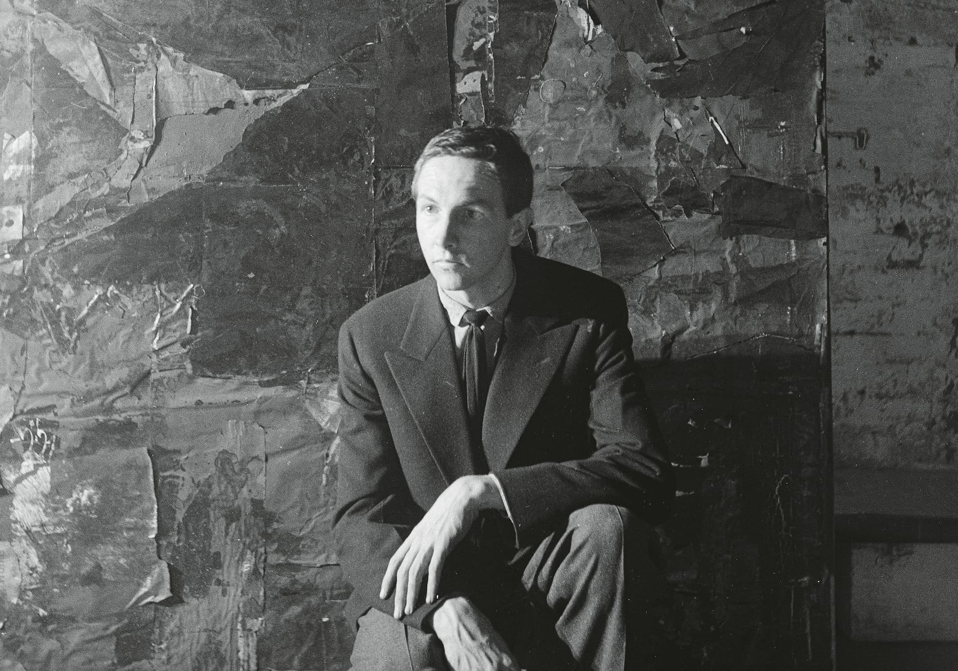 A man in a suit sits against a textured, abstract wall, with soft lighting highlighting his contemplative expression.