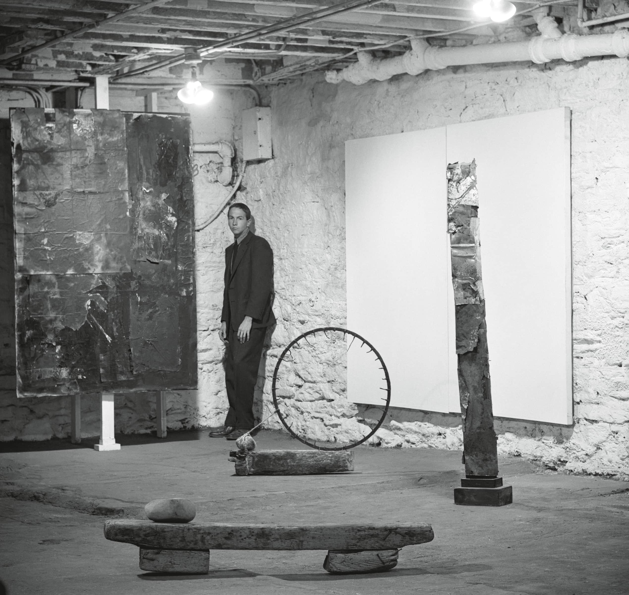 A man stands in a dimly lit basement gallery surrounded by abstract sculptures and large canvases on the walls.