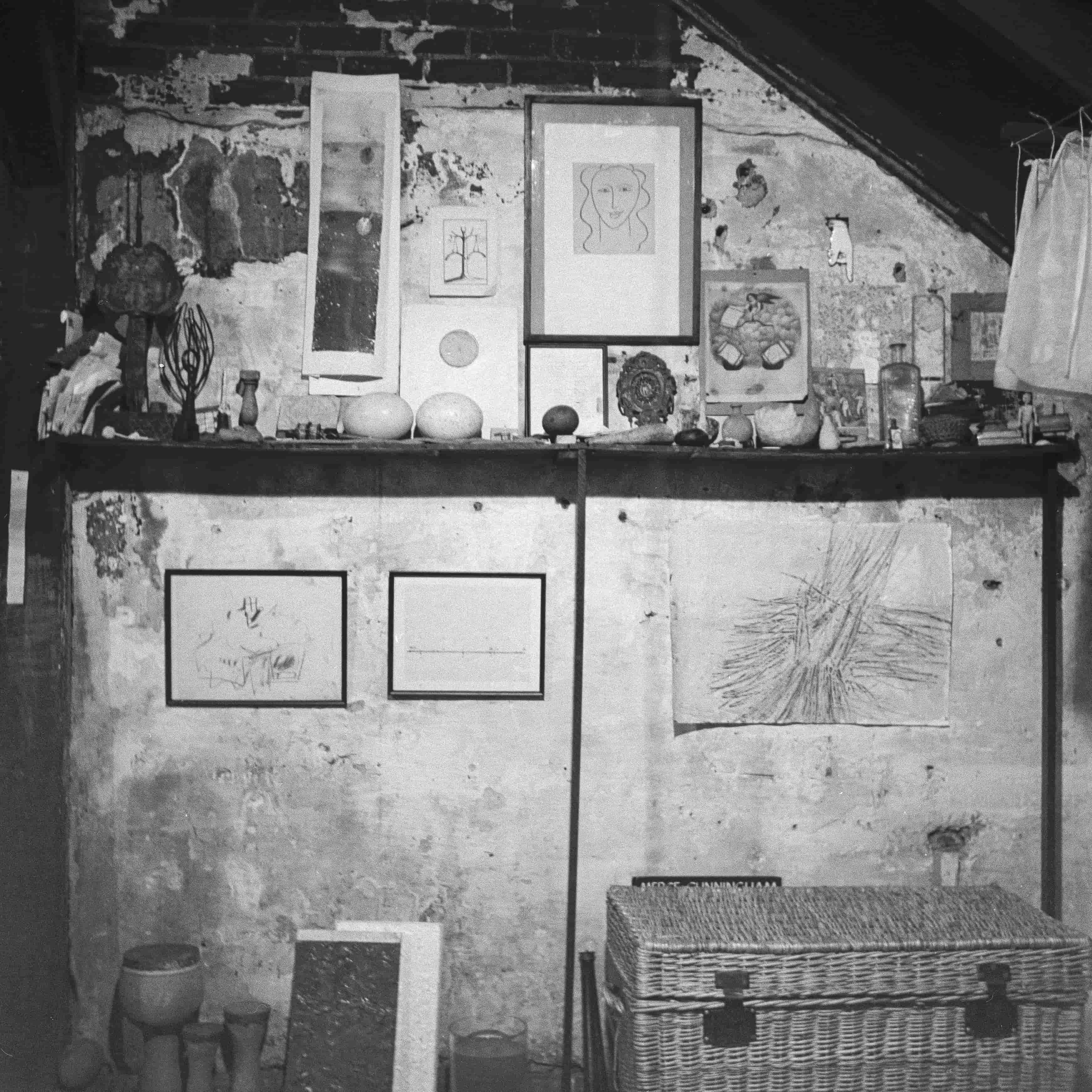 A black and white photograph of a rustic shelf against a textured wall, displaying various framed artworks, decorative objects, and a woven basket.