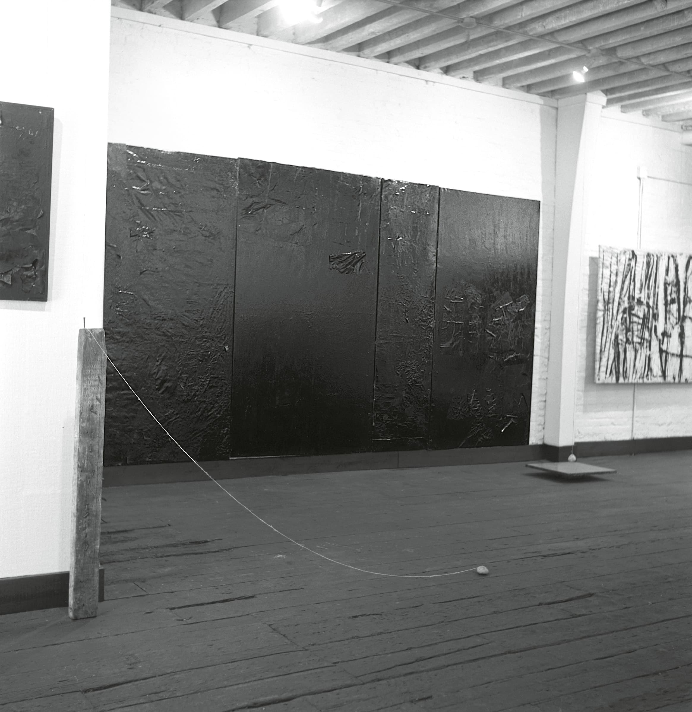 A black and white photograph of an art gallery featuring large black abstract paintings on the walls, wooden flooring, and a minimalist installation with a string and a small object on the ground.