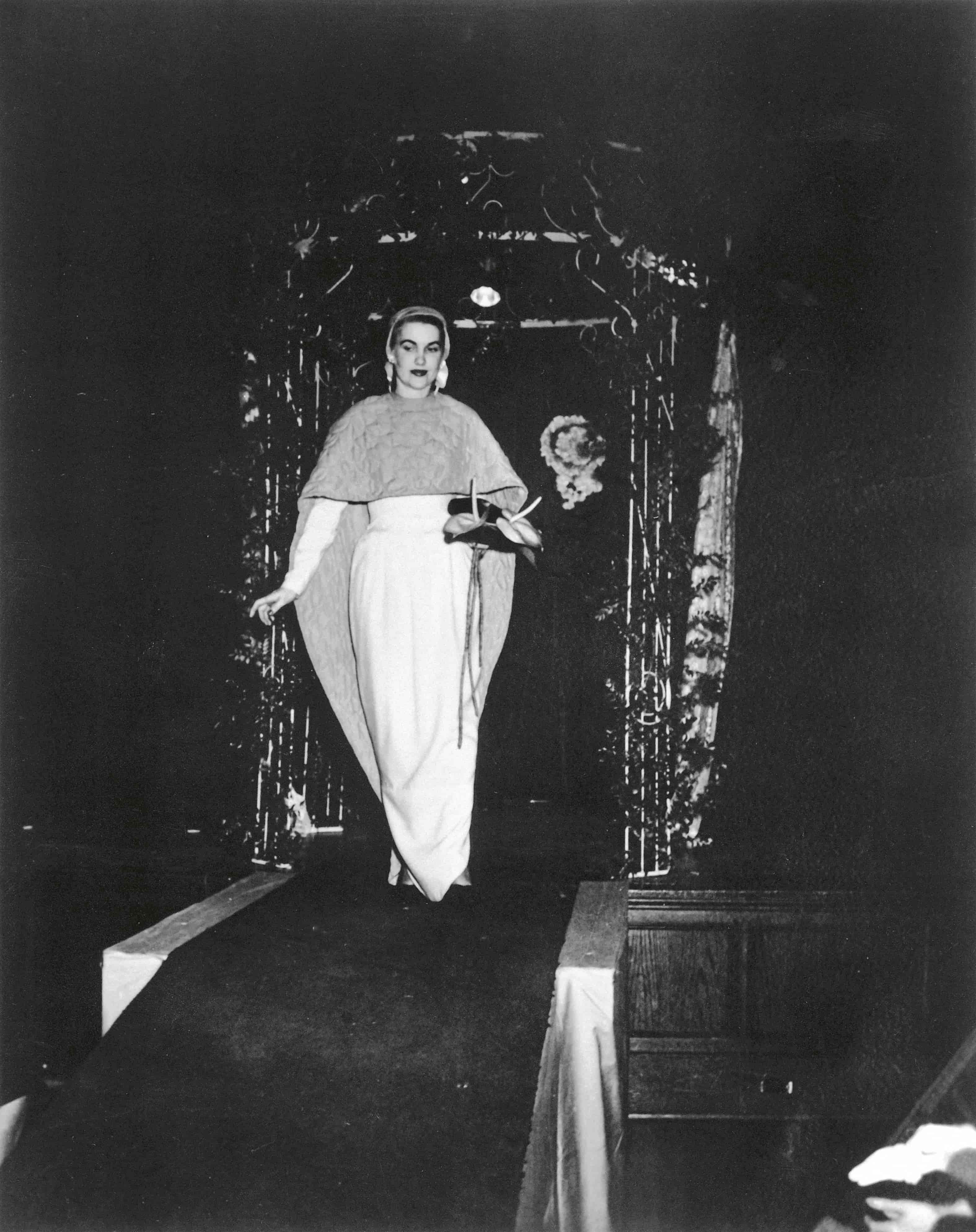 A woman in an elegant gown walks down a dimly lit runway, framed by decorative arches.