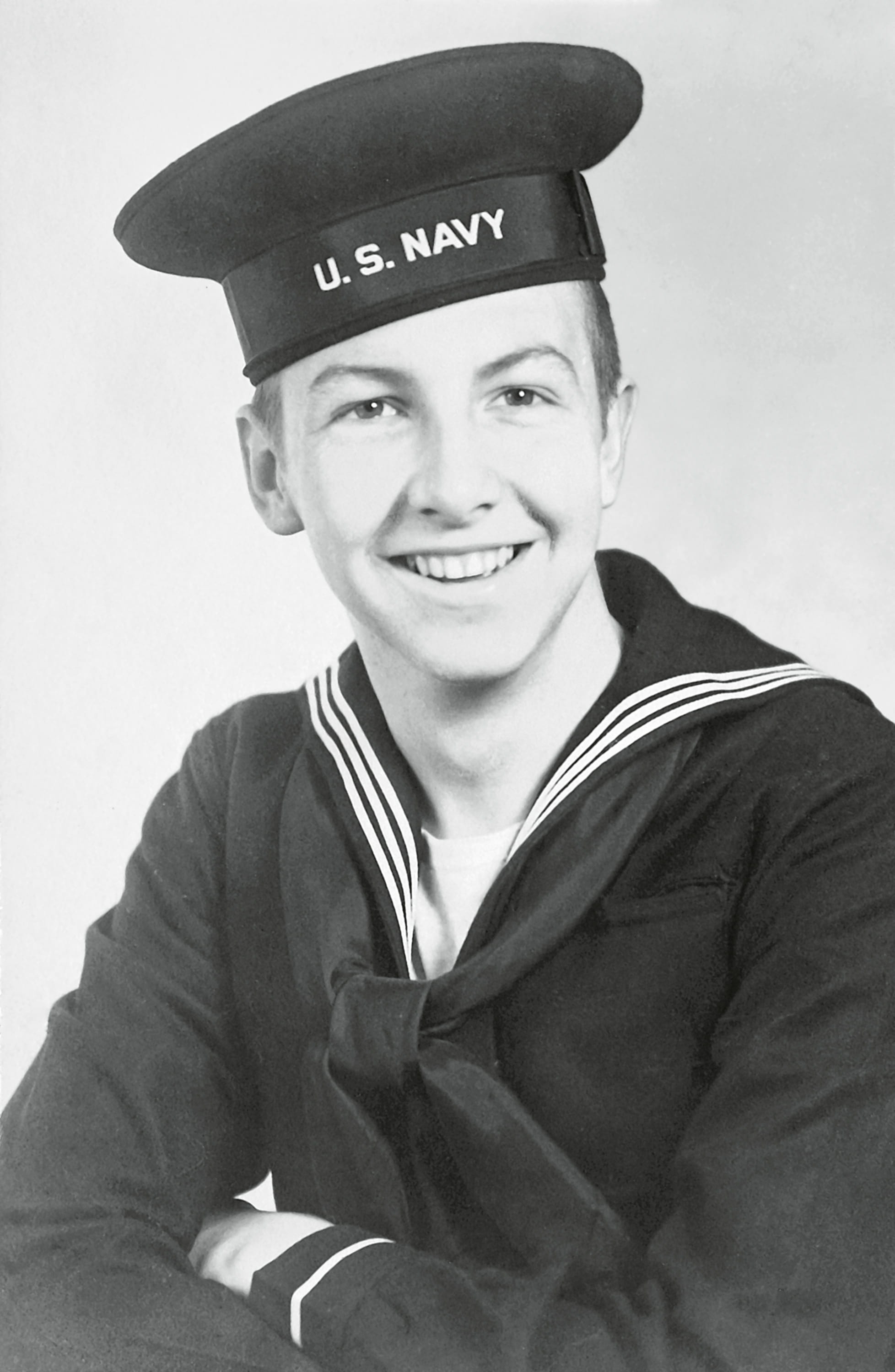 A young man in a U.S. Navy uniform, smiling and posing for a portrait with a sailor hat and a dark navy jacket.