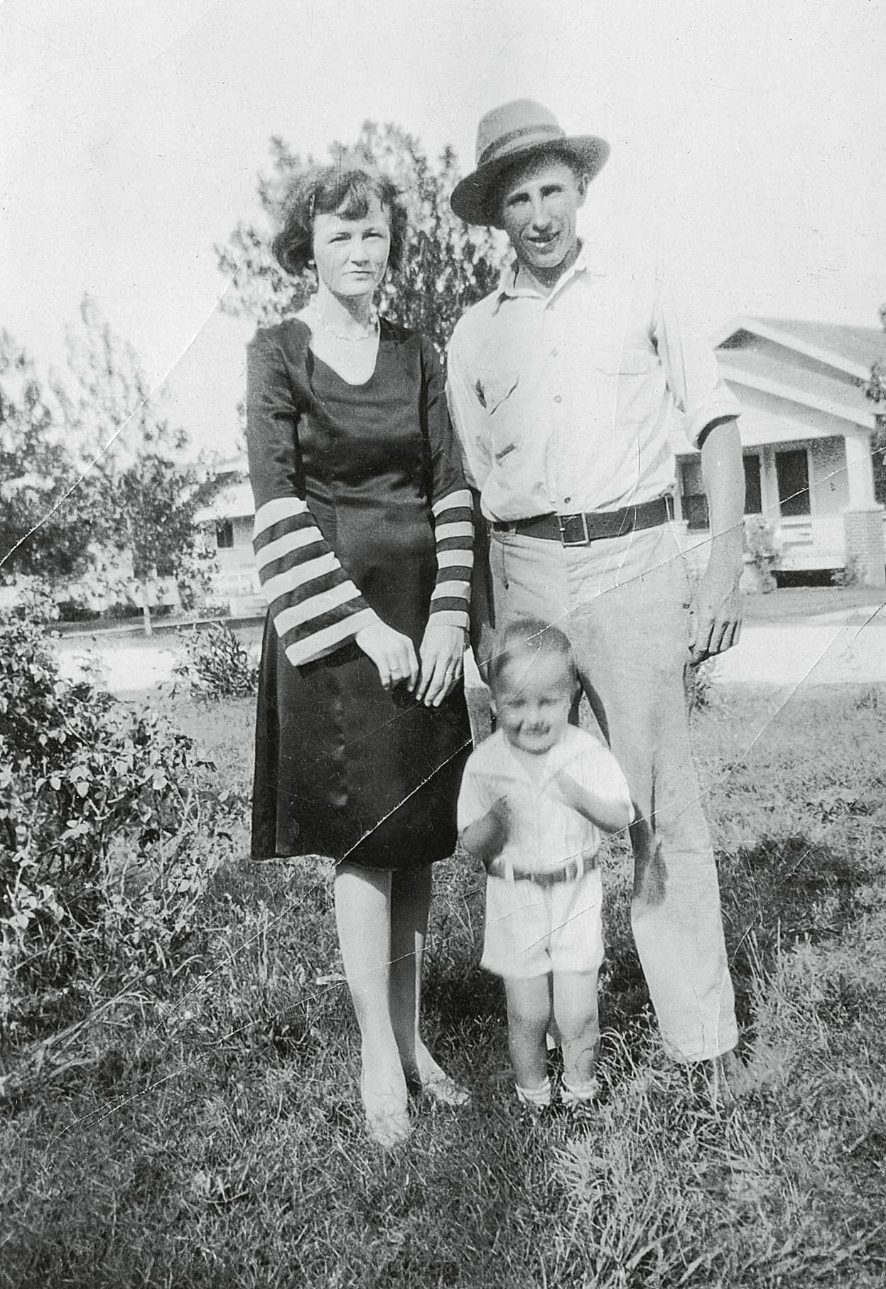 A black and white photograph depicts a family – a woman, a man, and a young boy – standing outdoors in front of a house.