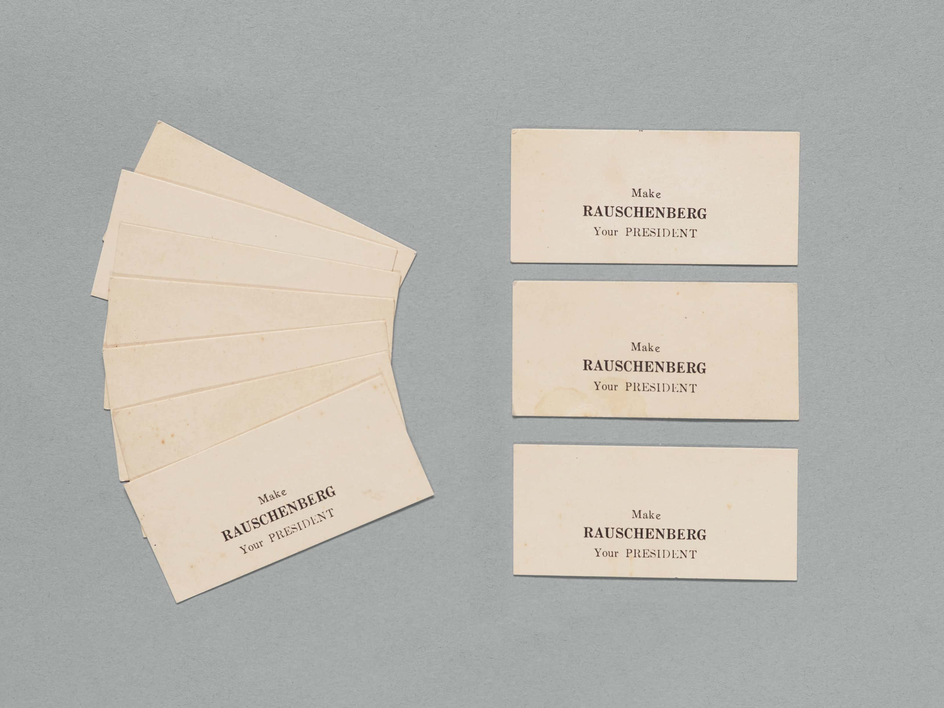 A collection of vintage cards with the text "Make Rauschenberg your PRESIDENT" printed in black on a light beige background, arranged in two groups on a gray surface.